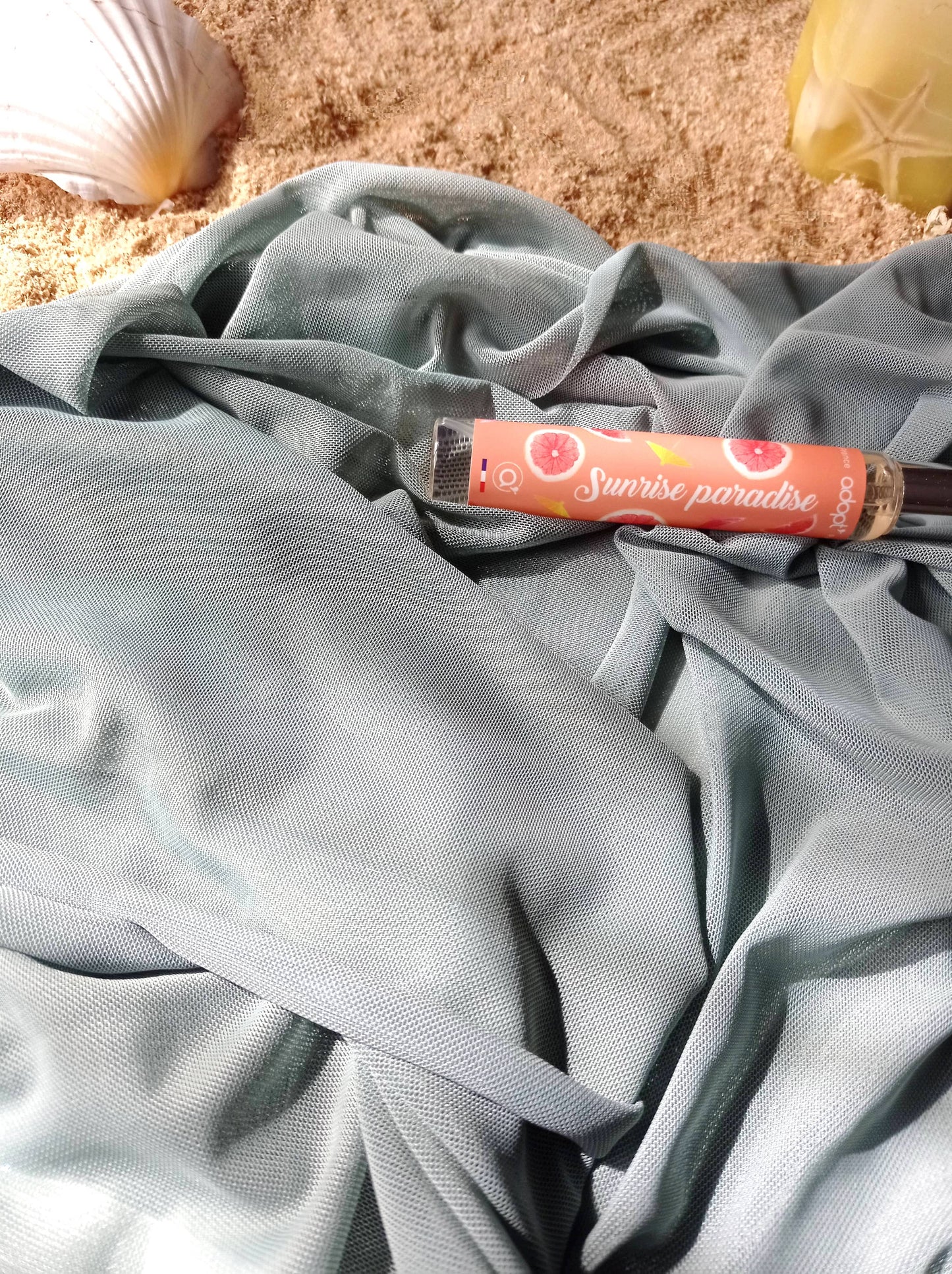 Tissu fin vert clair posé sur du sable entourée d'un coquillage et d'un parfum